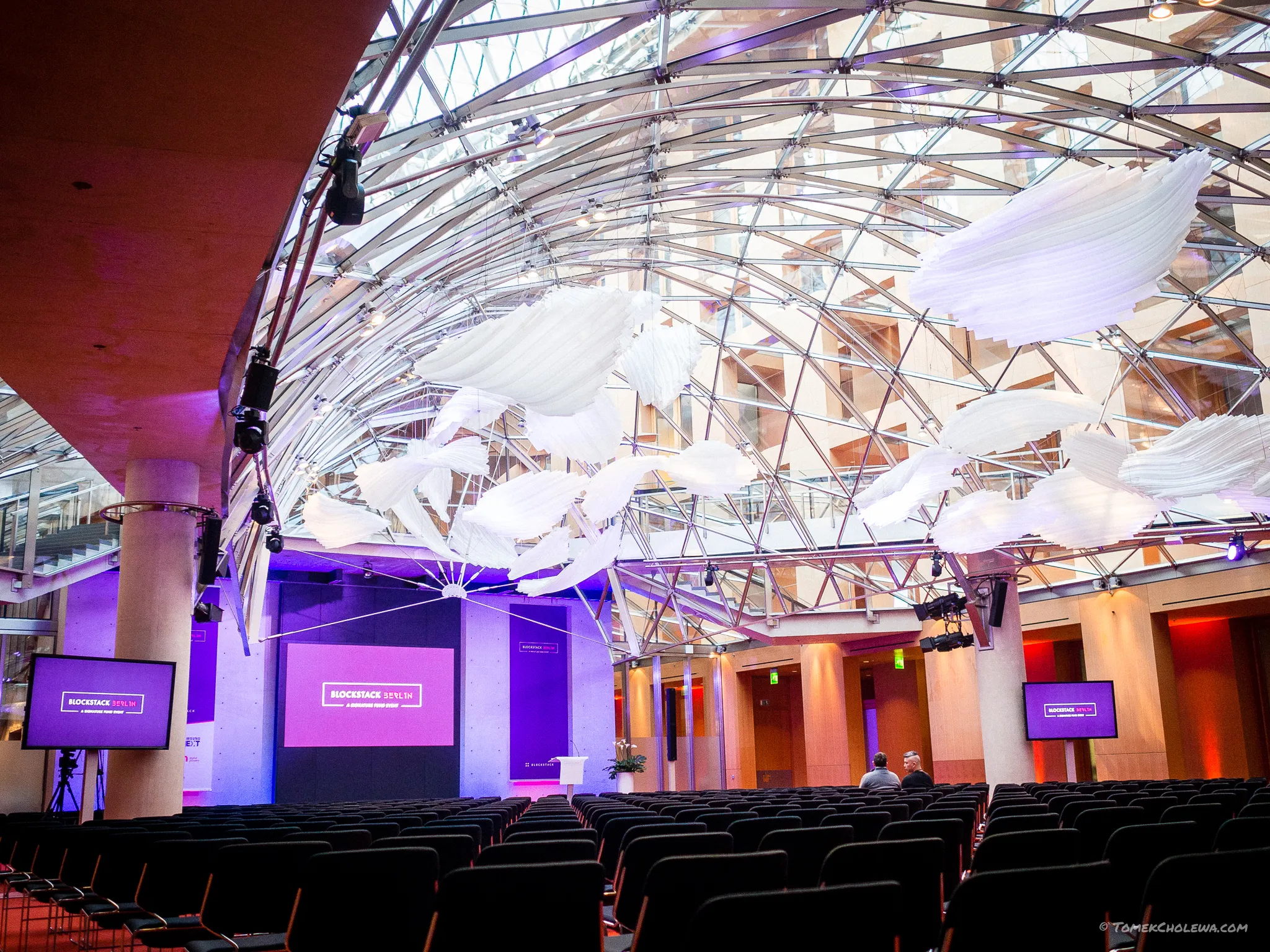 Empty auditorium beneath a suspended white installation and purple stage lights.