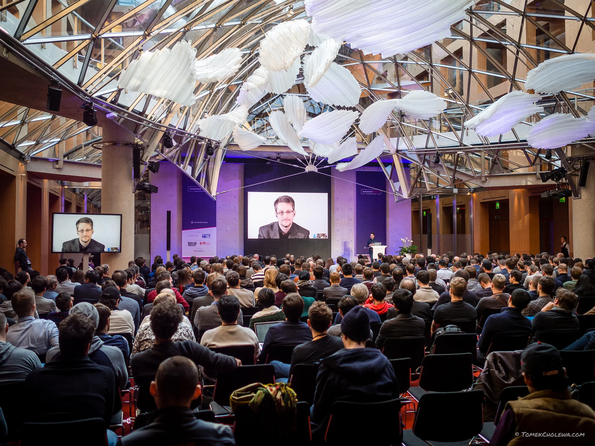 A packed auditorium watches a remote speaker on large screens.