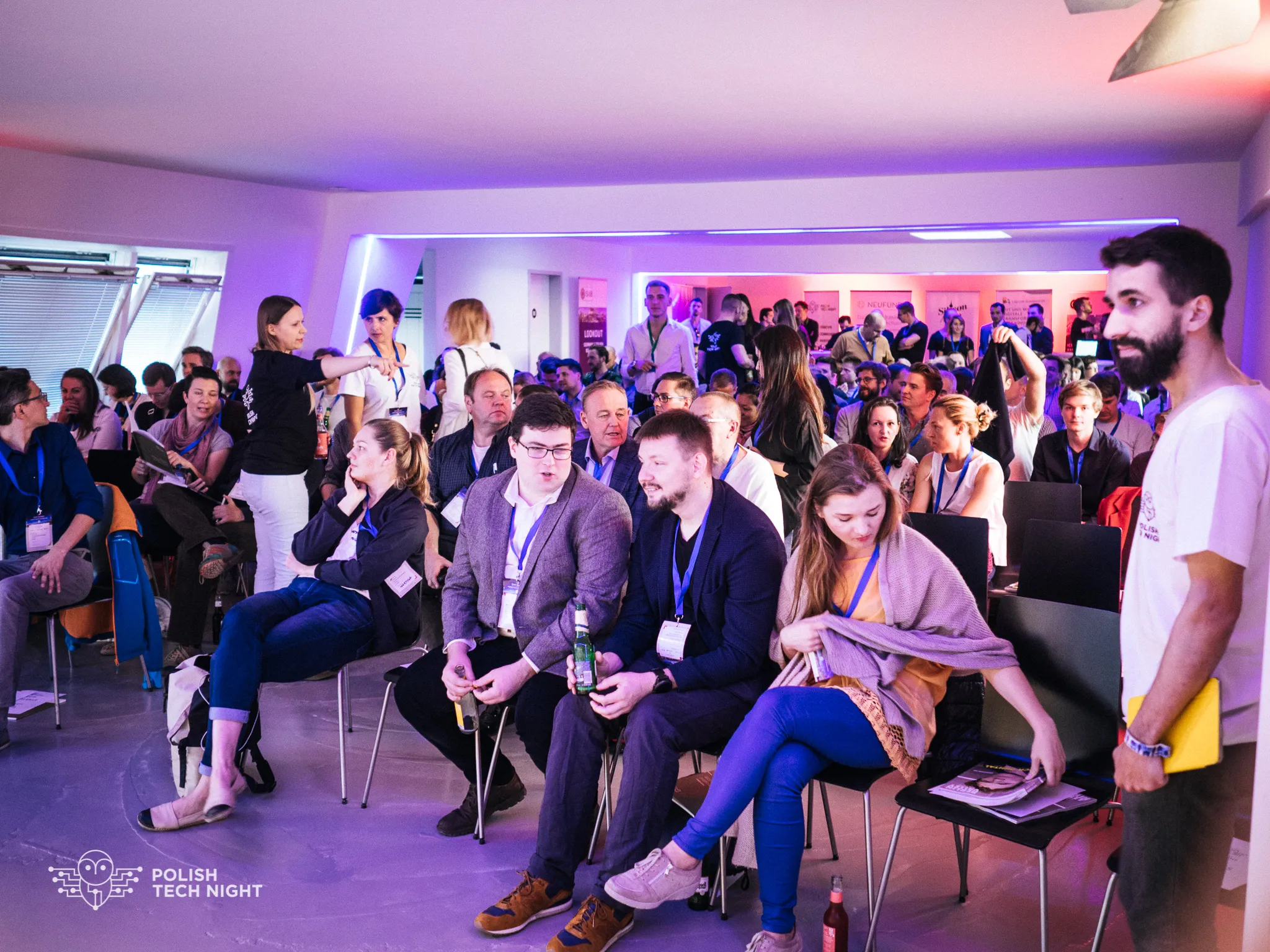 Attendees fill the room and talk before a session under colorful lighting.