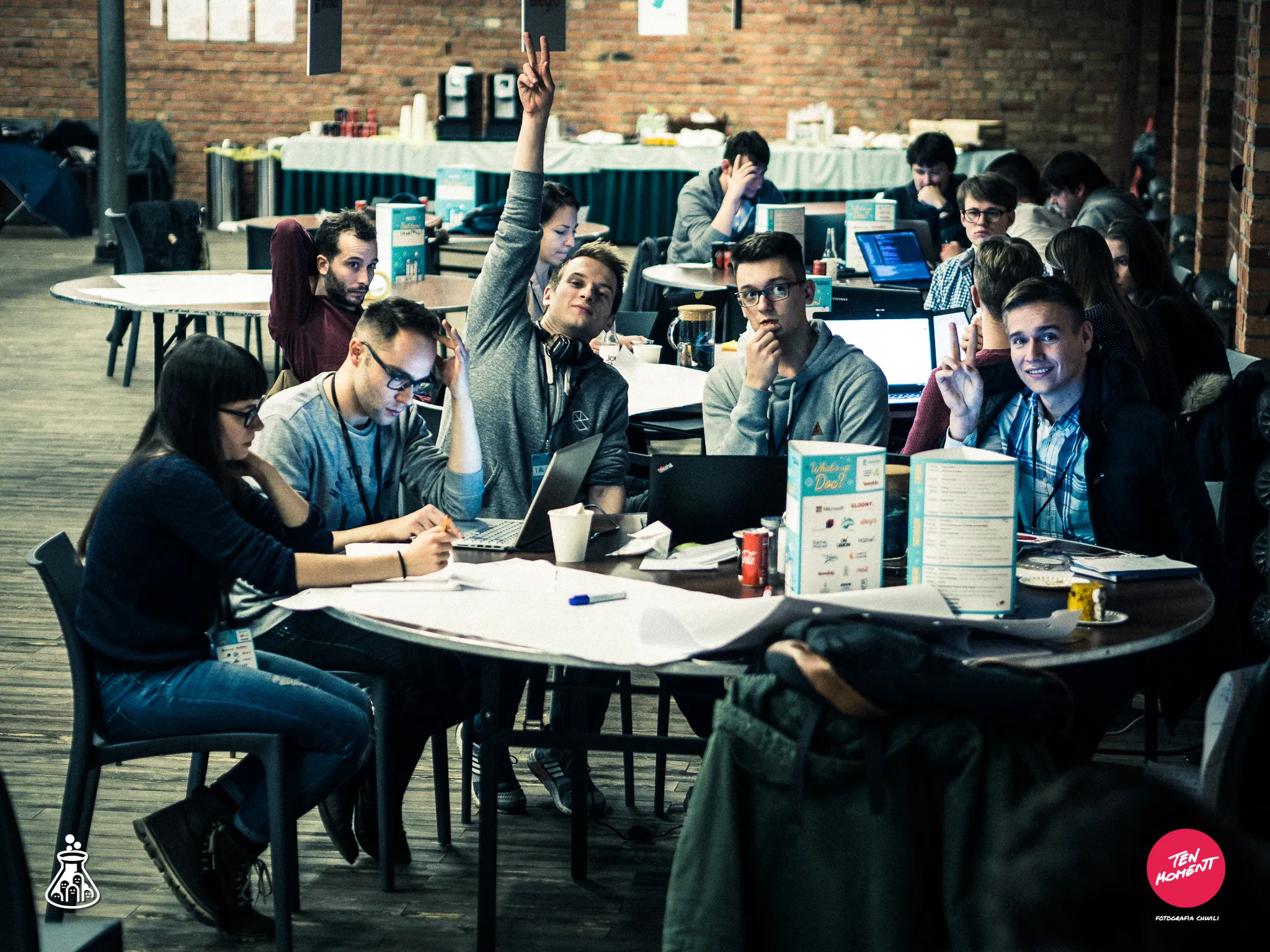 Hackathon participants work at tables with laptops while two pose with peace signs.