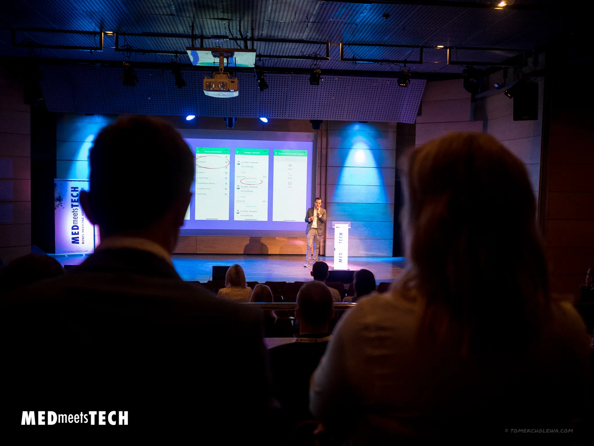 A presenter demonstrates a mobile app on a blue-lit stage before an audience.