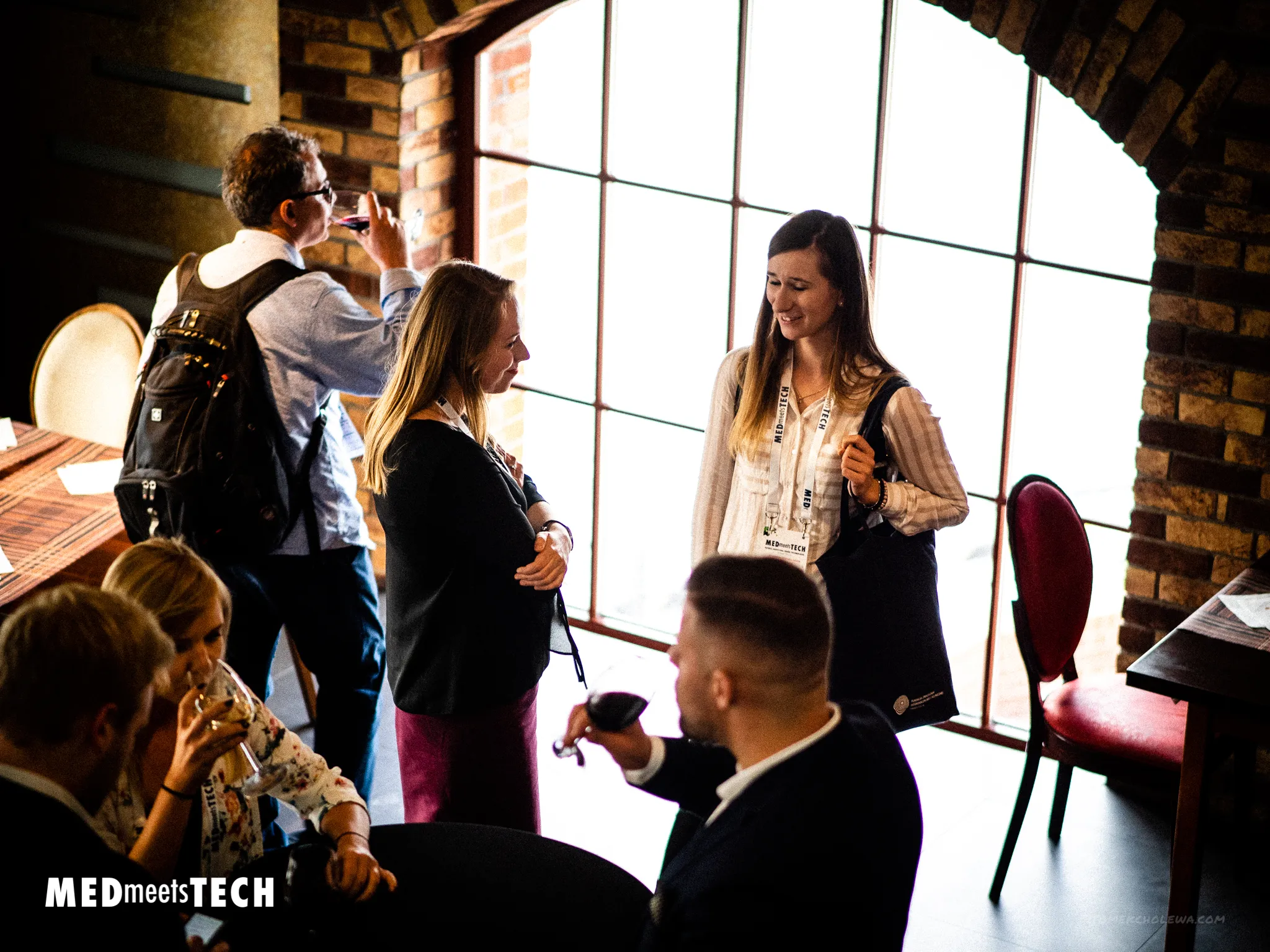 Attendees chat beside a bright window while holding drinks.