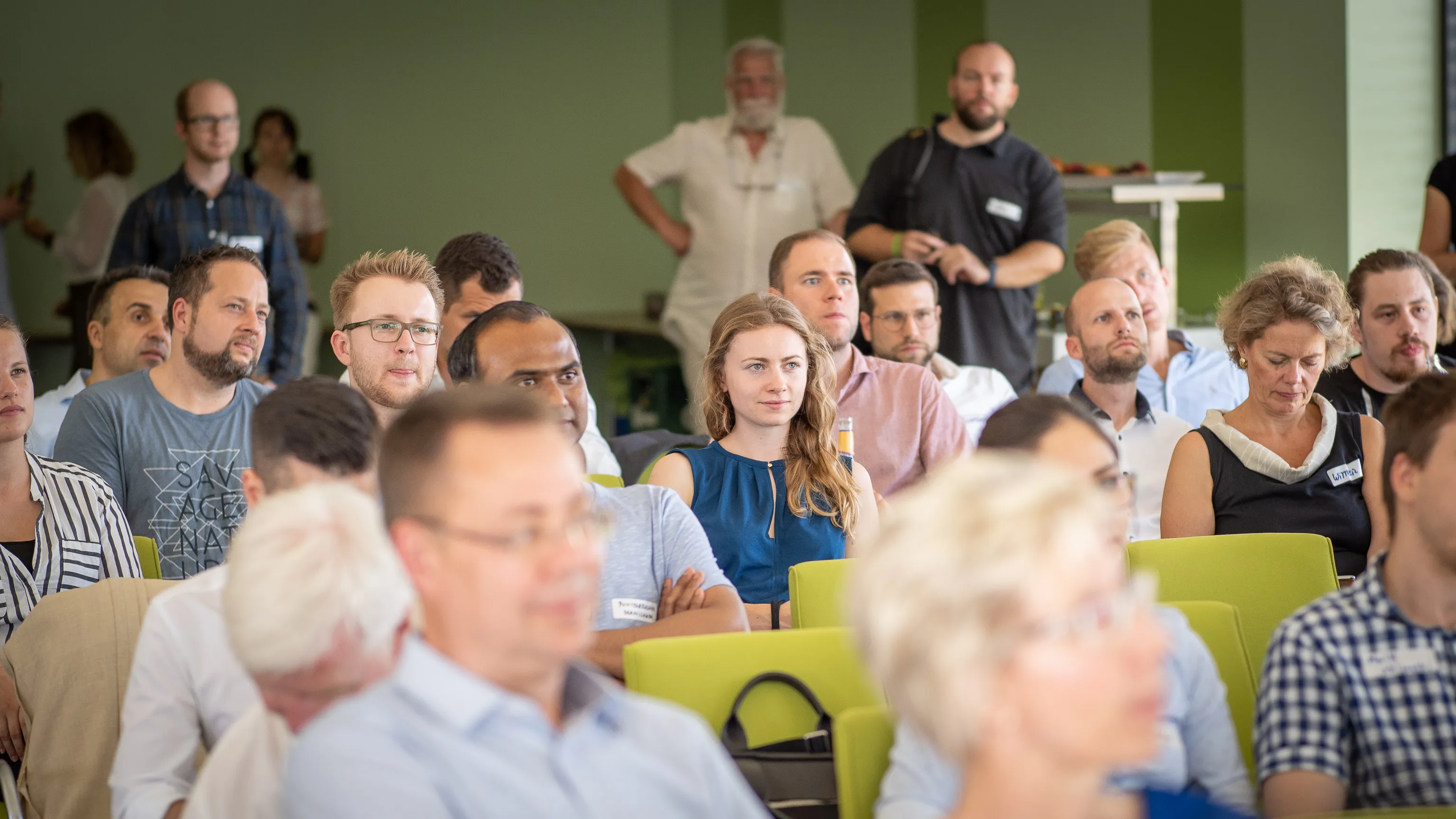 A focused audience fills rows of green chairs during a presentation.