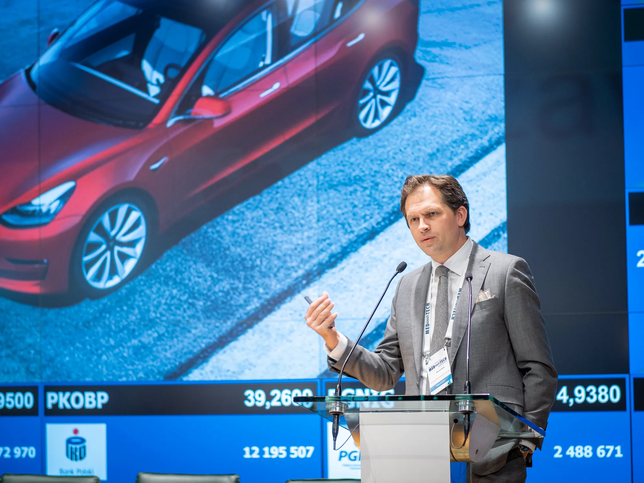 A suited presenter speaks at a lectern in front of a large image of a red car.