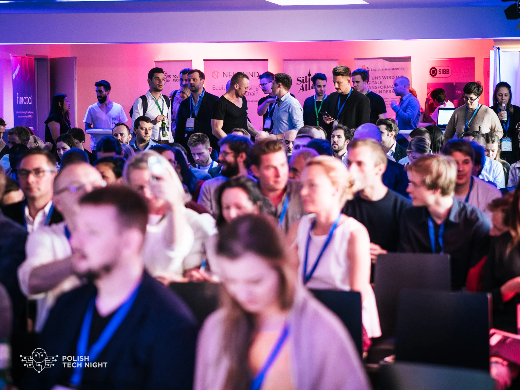A dense audience gathers beneath pink and blue lights with sponsor banners behind.