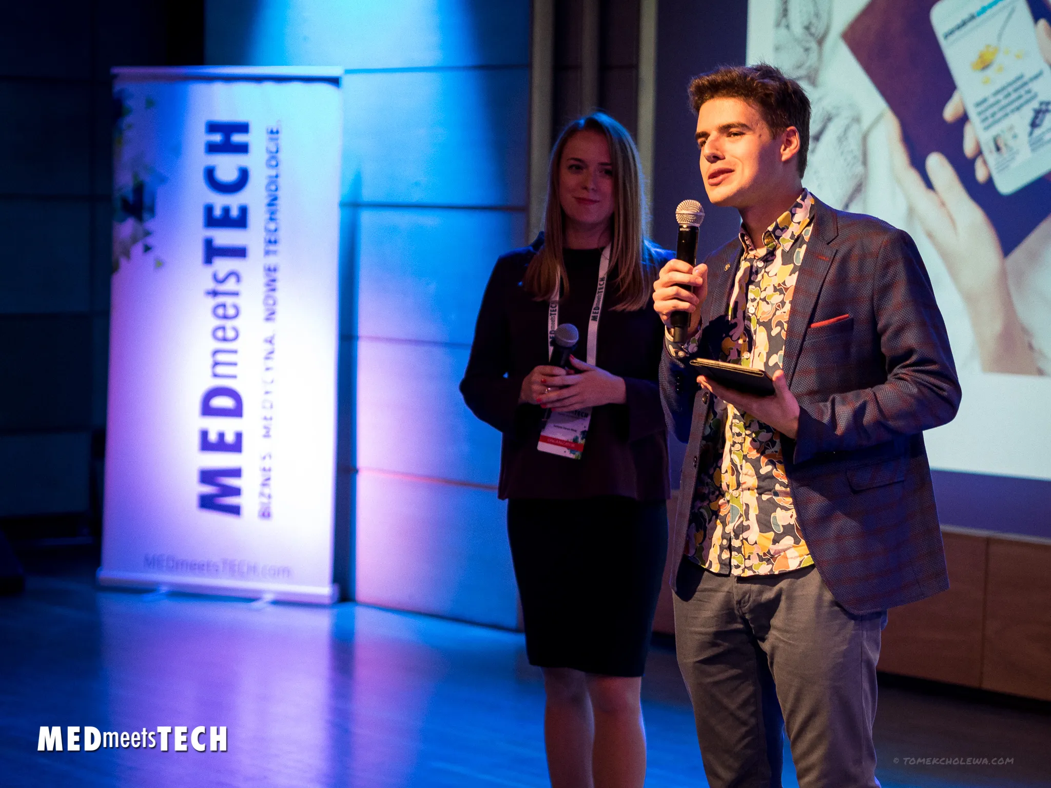 Two hosts speak into microphones on the blue-lit Med Meets Tech stage.