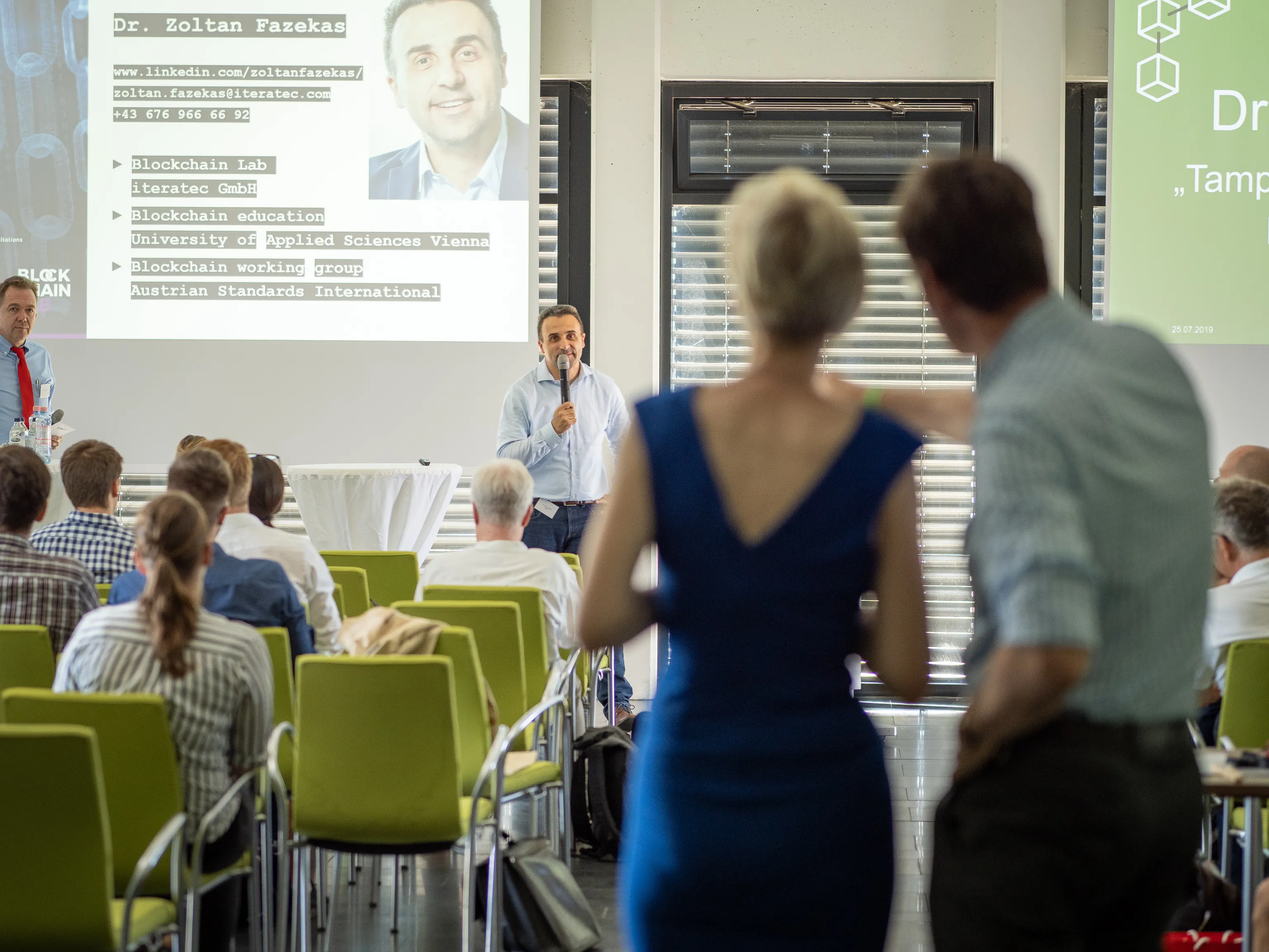 A speaker with a microphone presents blockchain topics as attendees cross the foreground.