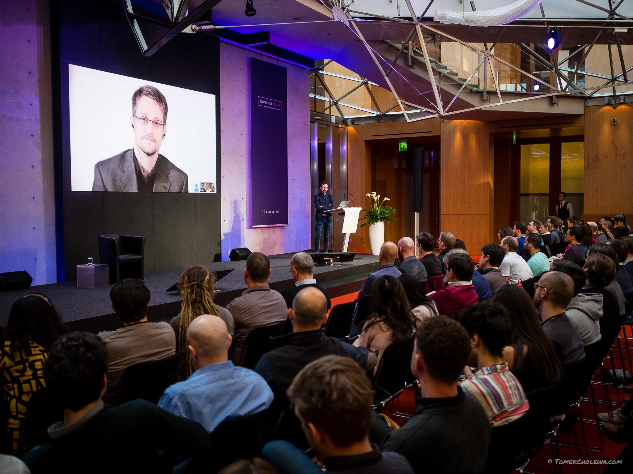 The audience listens to a presenter at a lectern with a remote guest onscreen.