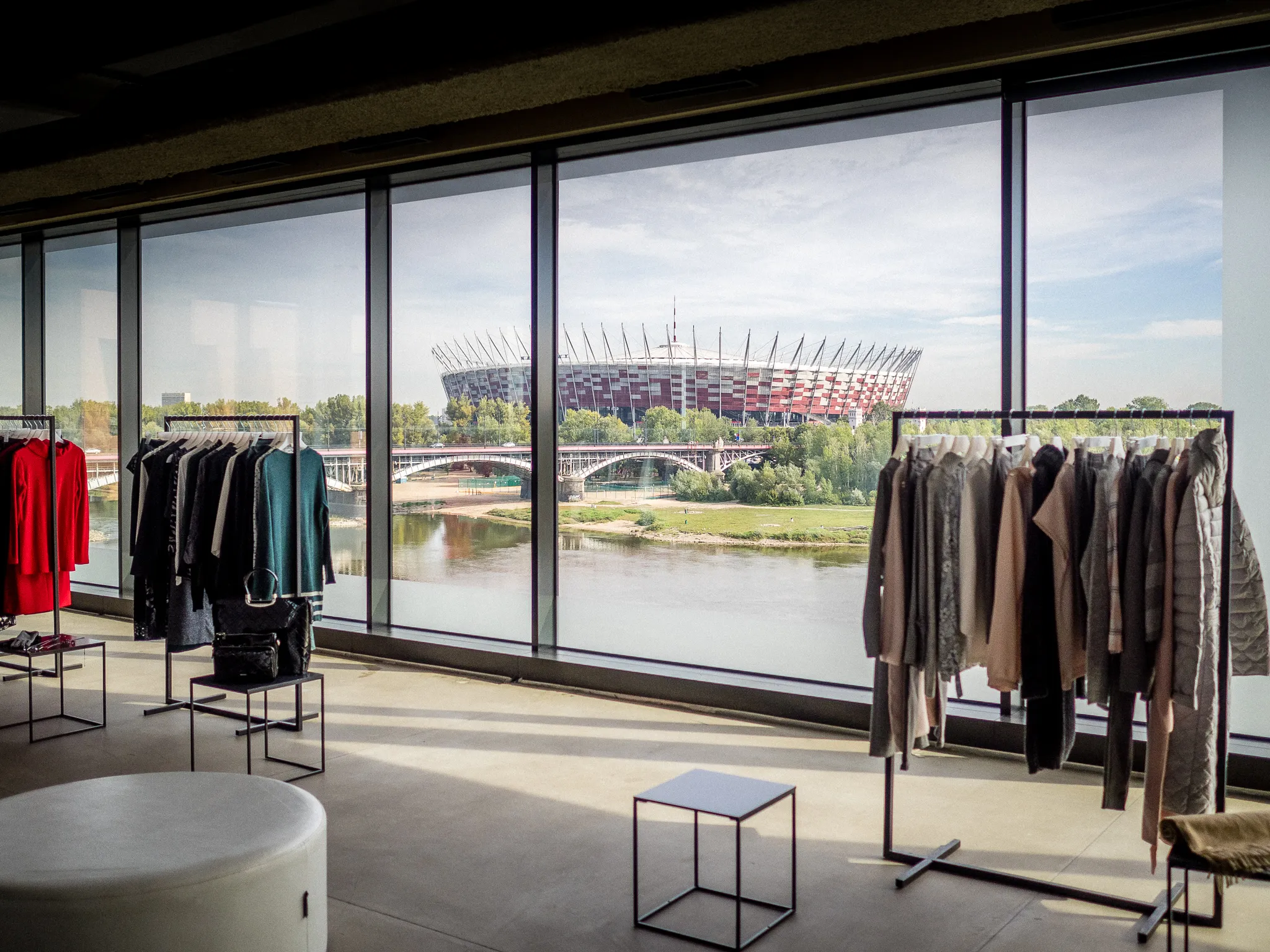 Clothing racks in a sunlit showroom with a stadium visible across the river.