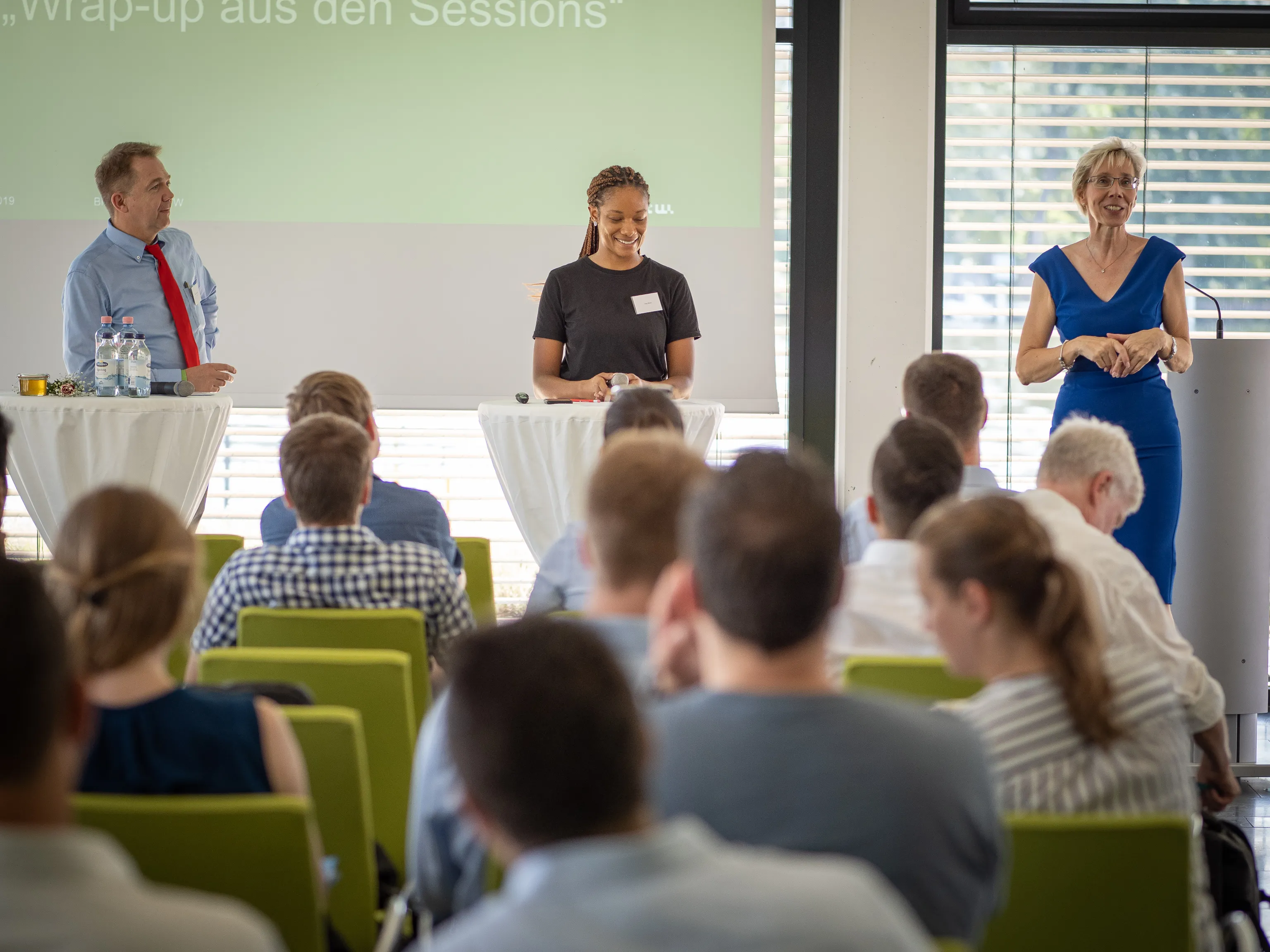 Three presenters stand before the audience during a session wrap-up.
