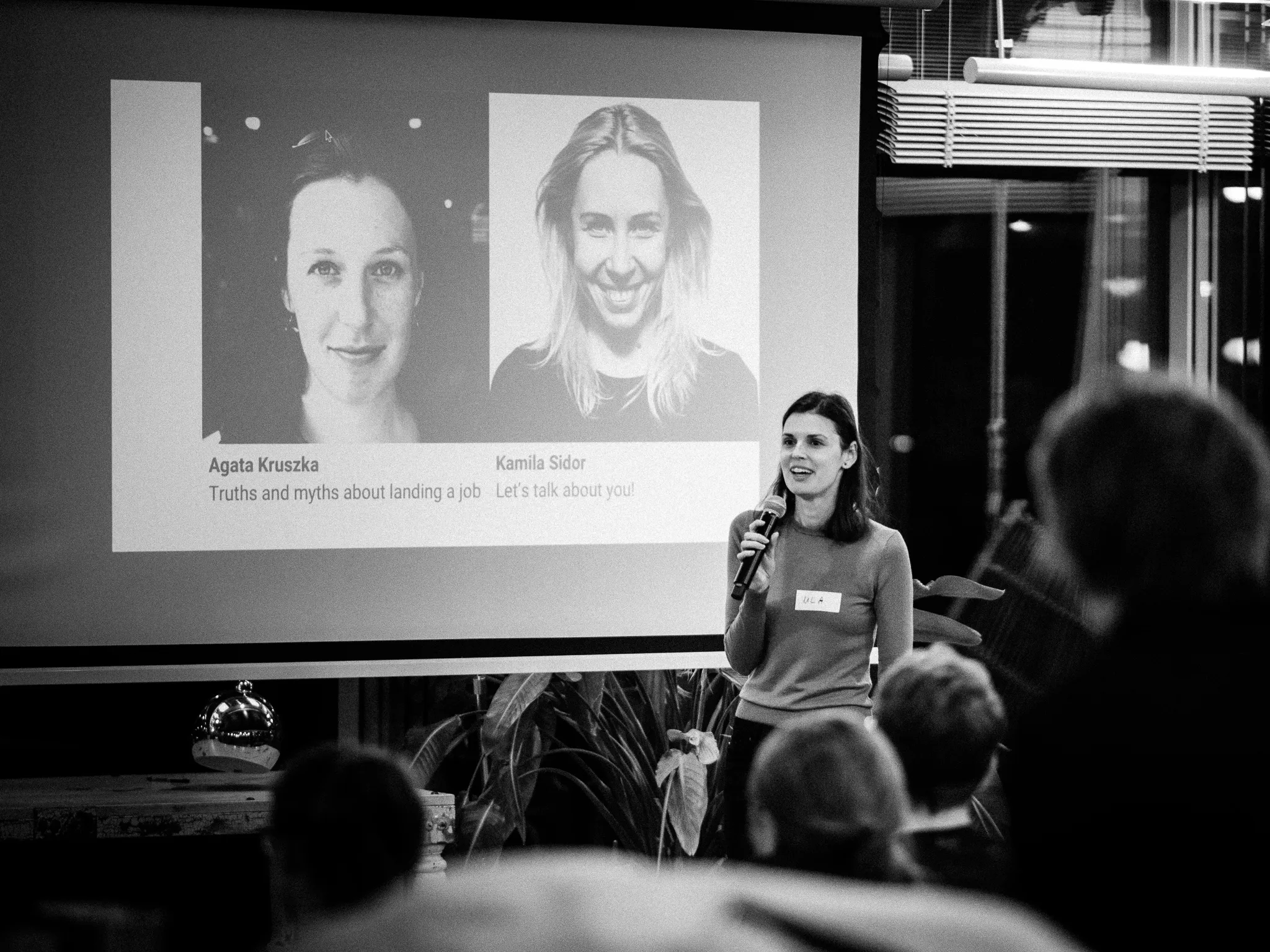 A woman presents with a microphone beside a projected slide in a black-and-white frame.