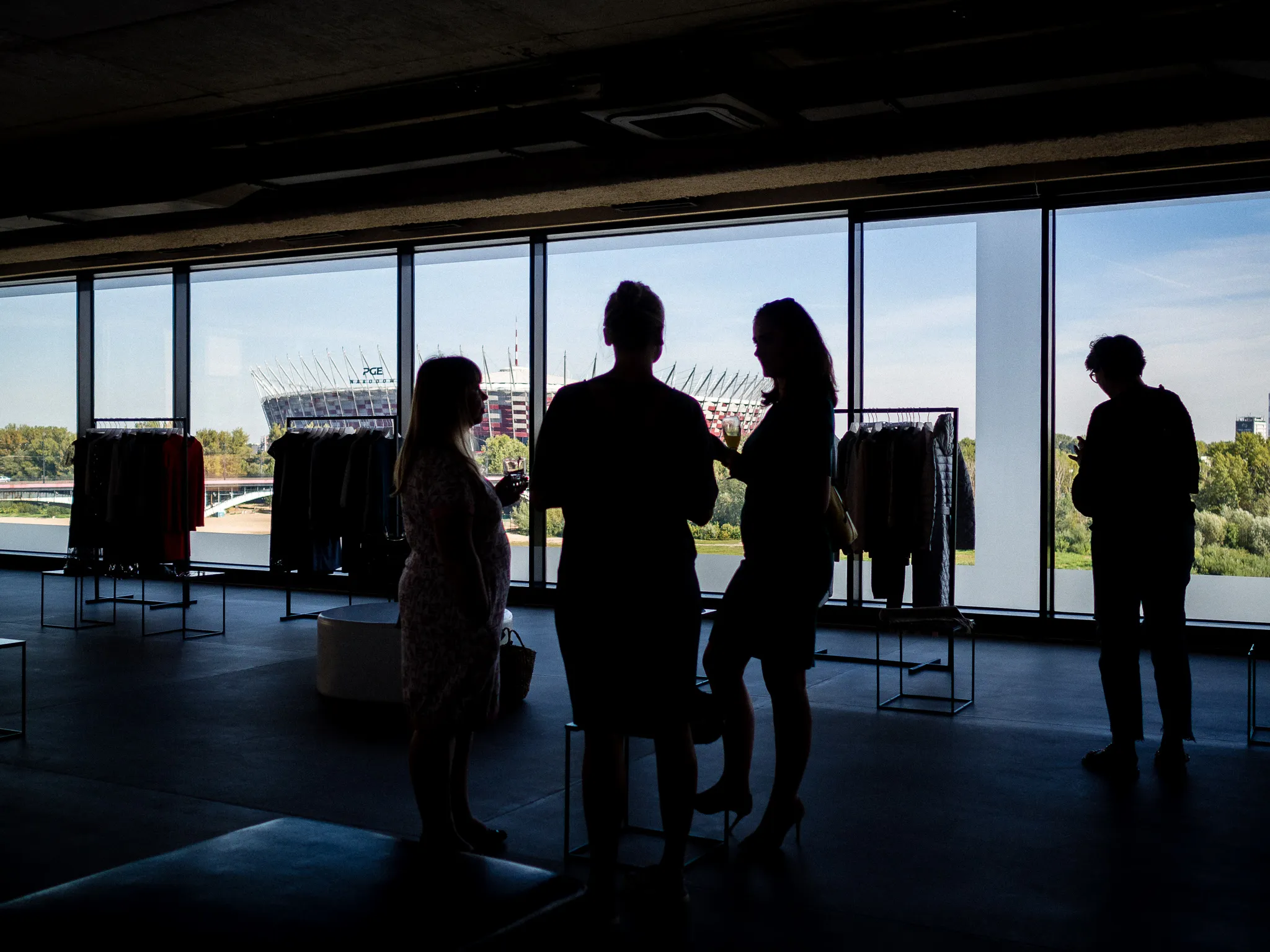 Silhouetted guests talk beside clothing racks and tall windows overlooking a stadium.