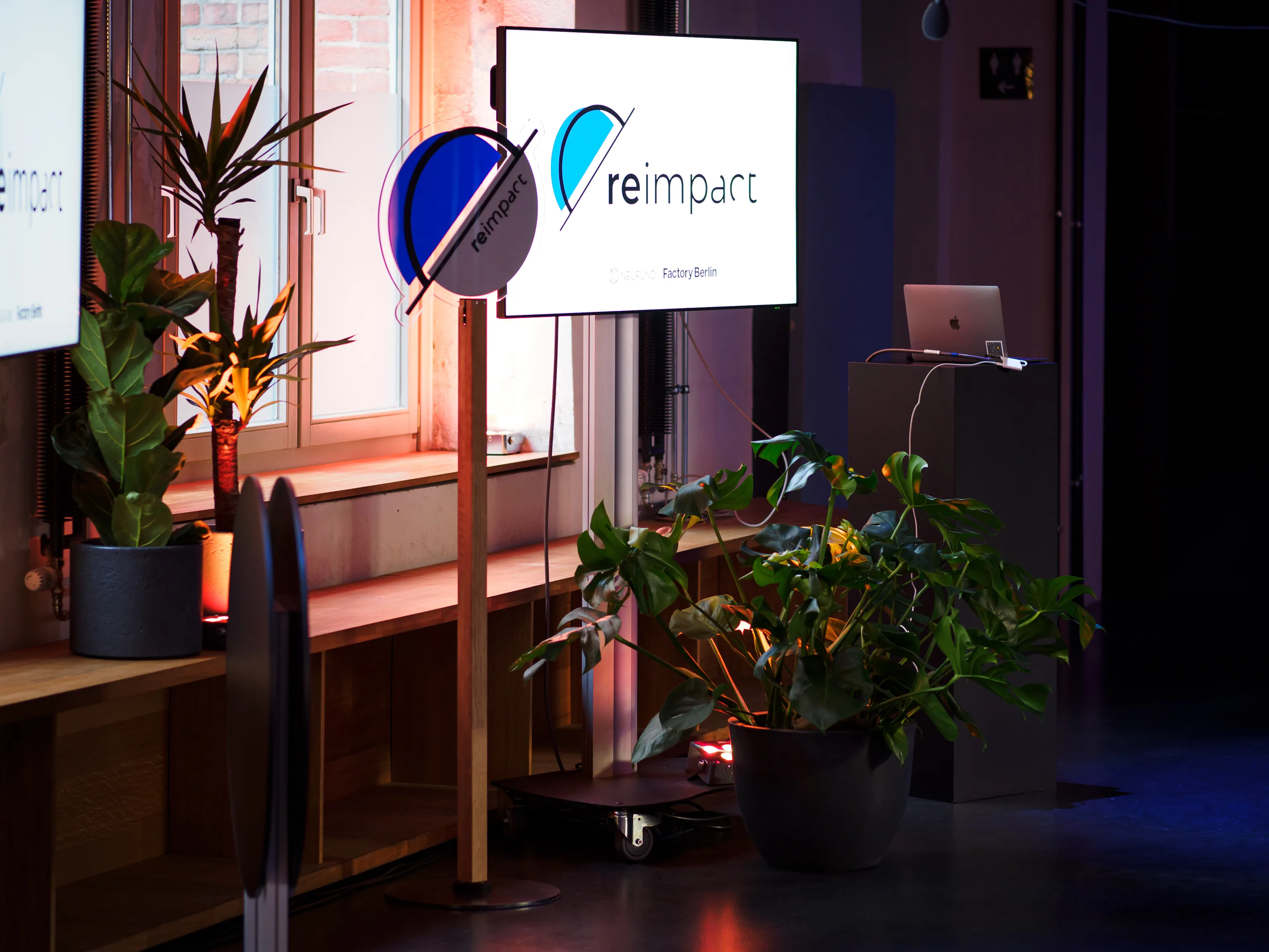Reimpact stage screens and a lectern surrounded by potted plants.