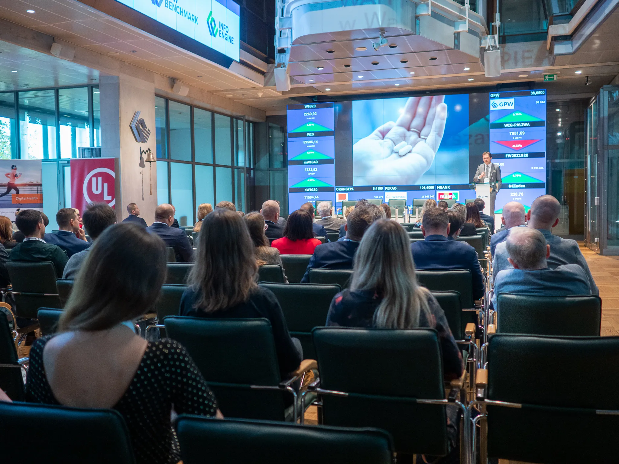 An audience watches a suited speaker as an image of medicine fills the screen behind him.