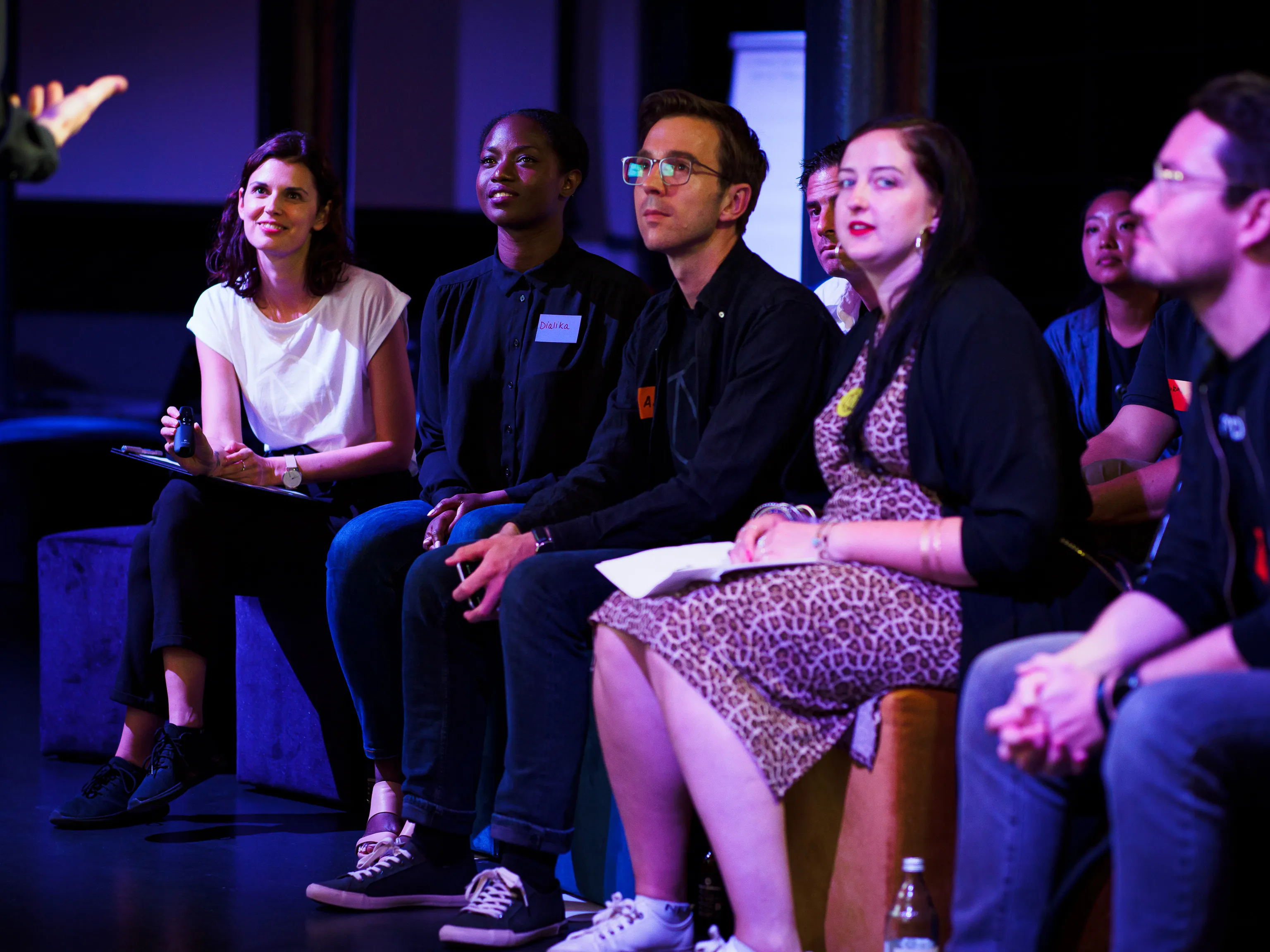 A seated workshop group follows a discussion under purple stage lighting.